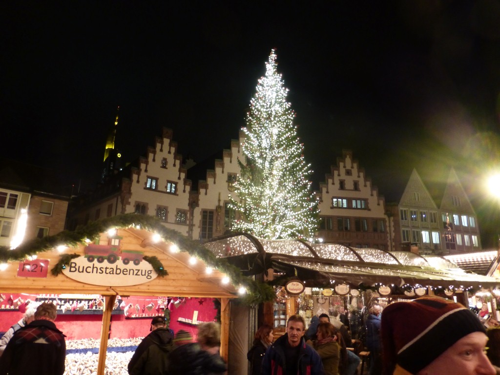 Frankfurter Weihnachtsmarkt mit Buchstaben-Eisenbahn-Stand, Tannenbaum und Römer Fassade im Hintergrund.